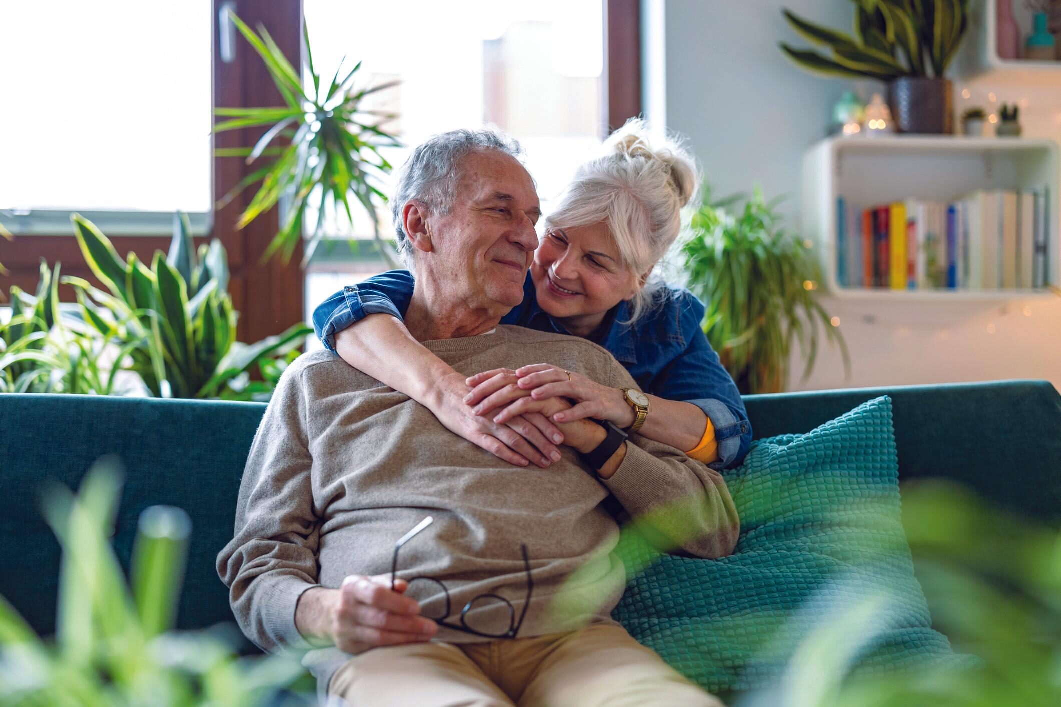 Older woman hugging older man from behind whilst he's sat on a sofa, green plant in the foreground