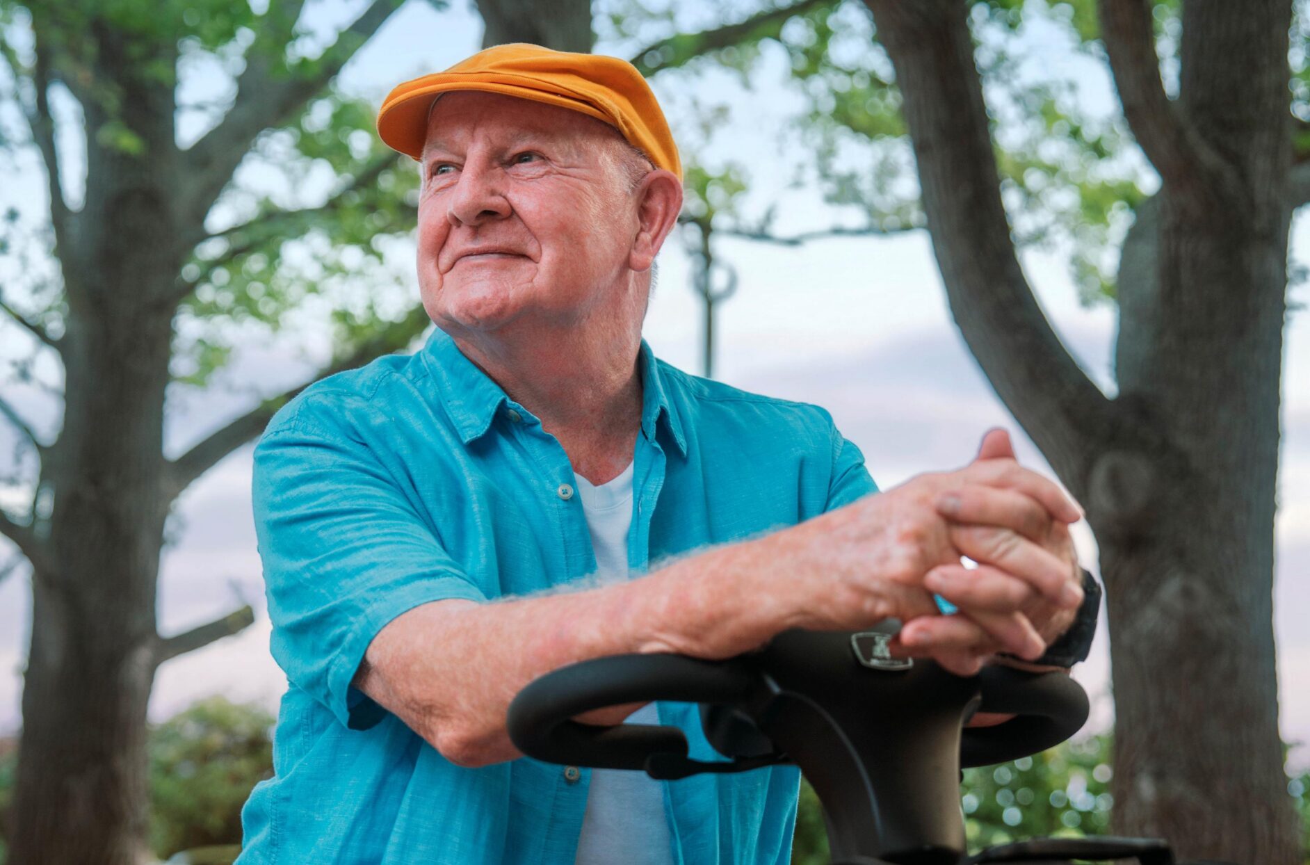 Close up of smiling older man wearing a yellow cap looking into the distance whilst sat on a charcoal Humfree connect scooter in the park