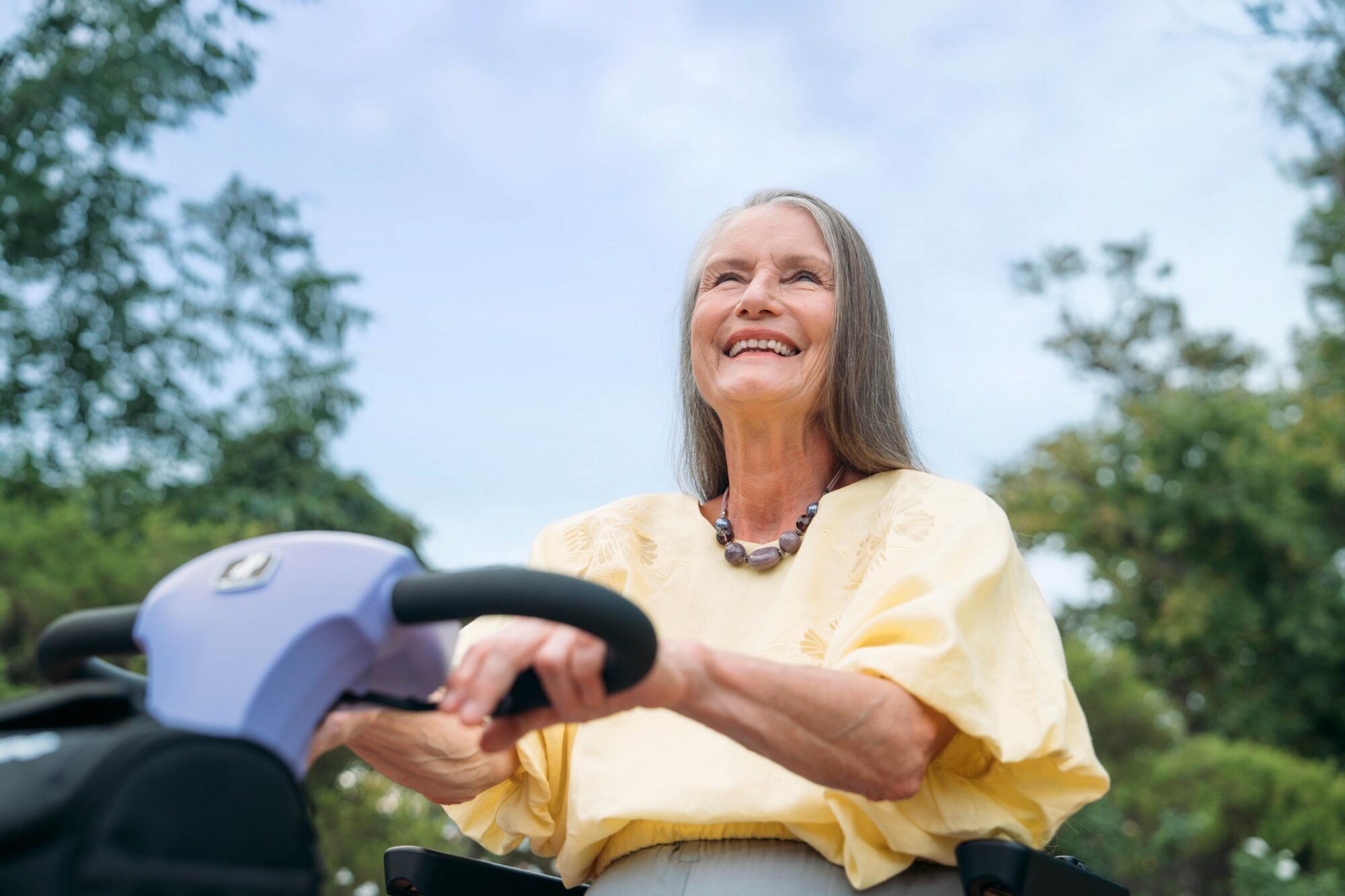 Close up of older woman wearing a yellow shirt smiling as she rides an amethyst Humfree connect scooter through the park