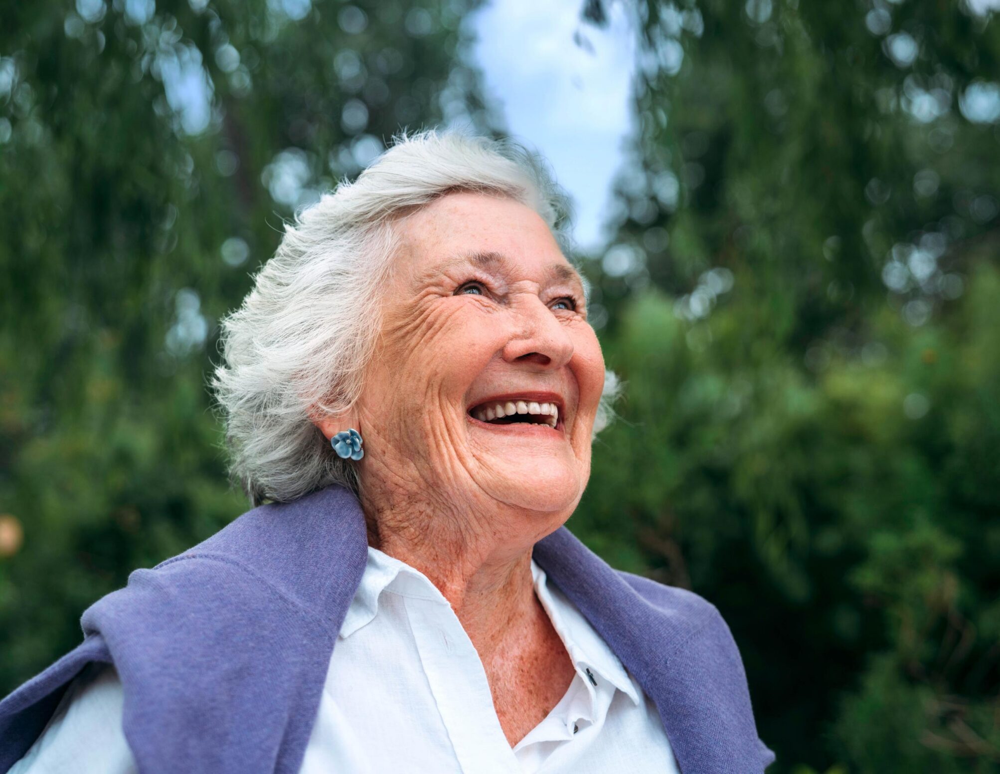 Close up of older woman in the park with a purple cardigan draped over her shoulders smiling into the distance
