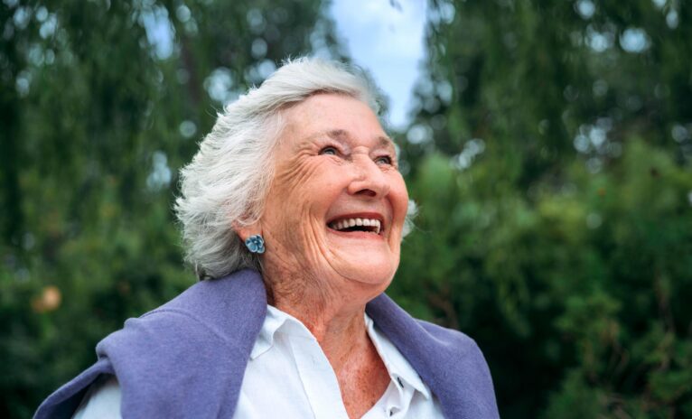 Close up of older woman in the park with a purple cardigan draped over her shoulders smiling into the distance
